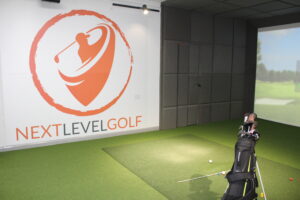 Image shows the inside of one of the Next Level Golf studio rooms, with the Trackman virtual golf screen, a golf bag with golf clubs and the Next Level Golf logo wall